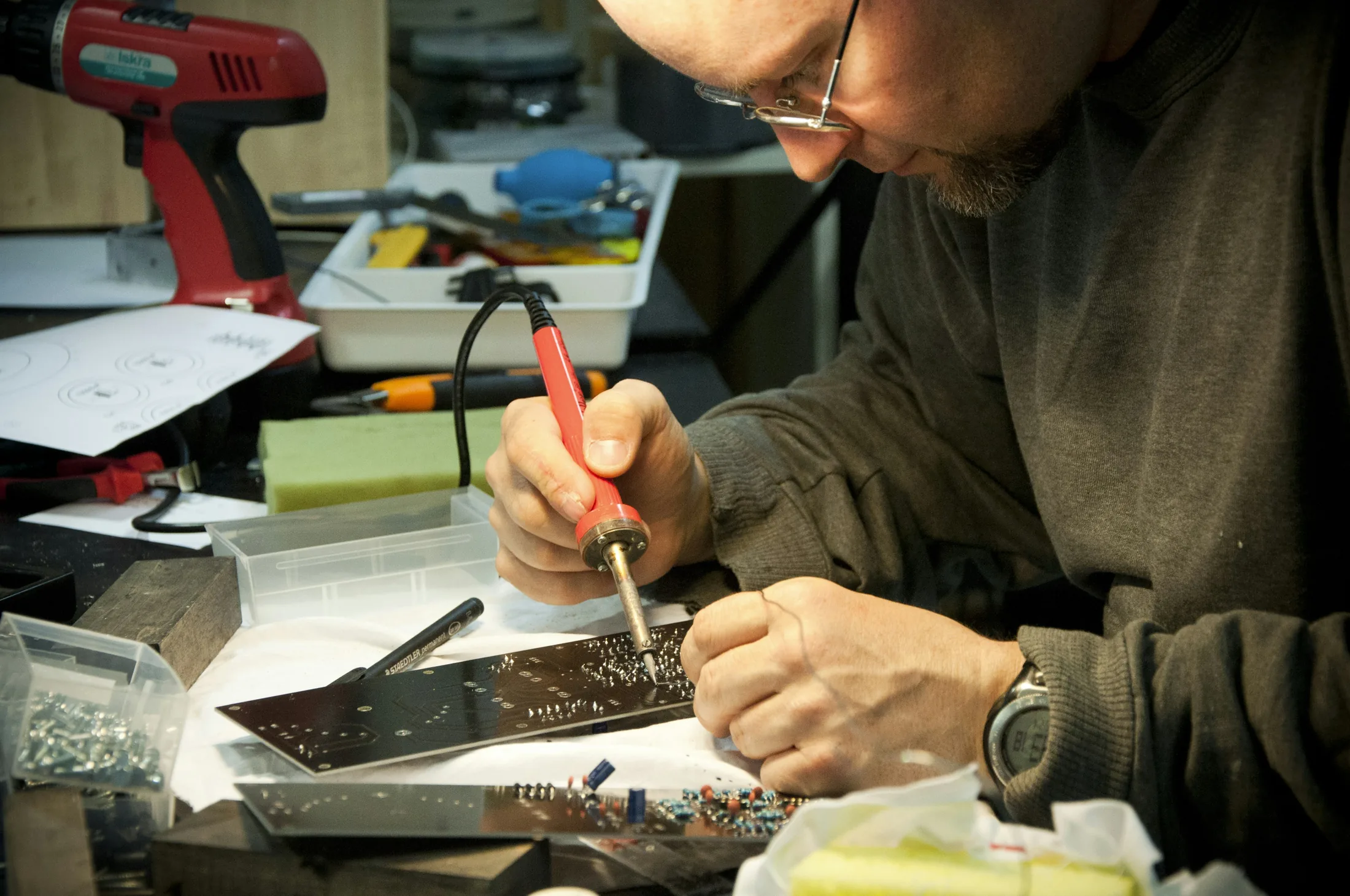 A person wearing glasses solders a circuit board at a workbench surrounded by tools and electronic components